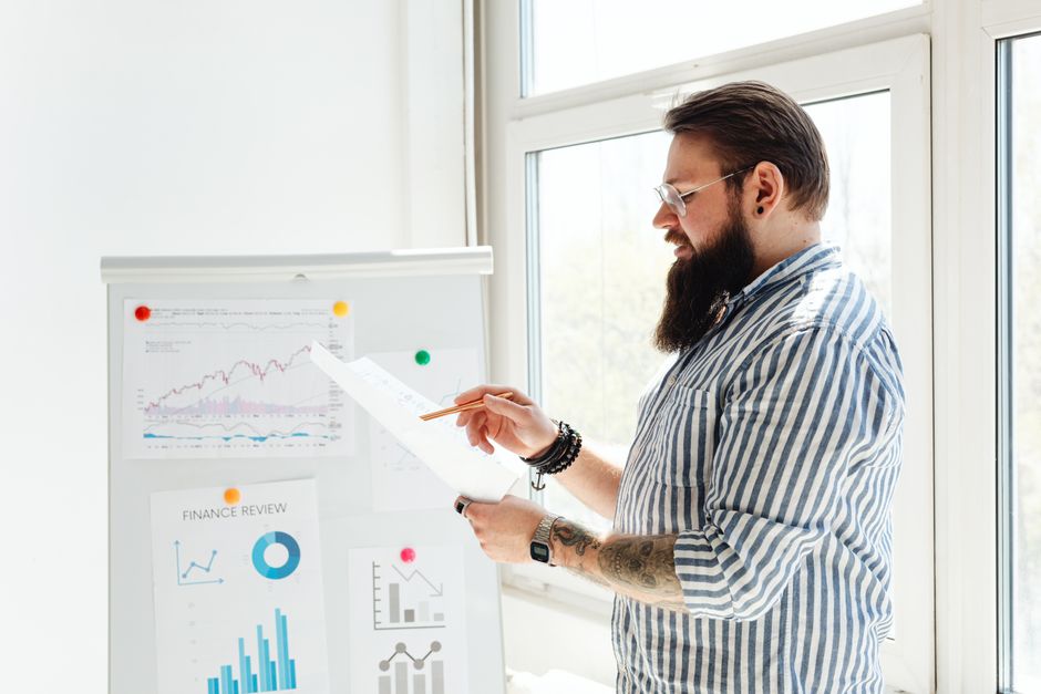 Um homem de barba e óculos analisa gráficos financeiros em papel ao lado de um quadro branco.